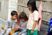 A child sits holding a tablet while another child sits and looks on and another stands looking on.