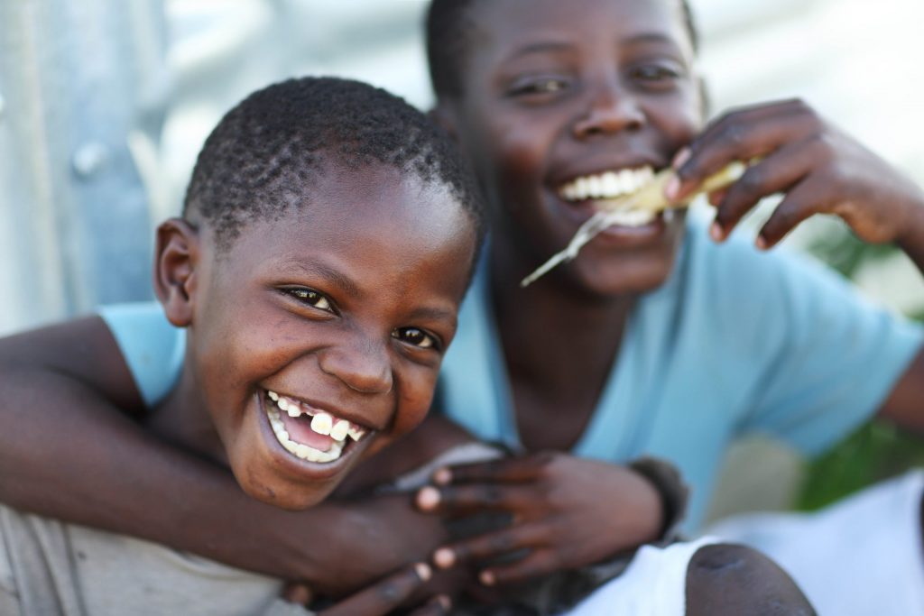 A photo of two children wearing blue shirts smiling, the one in back has their arms around the one in front.
